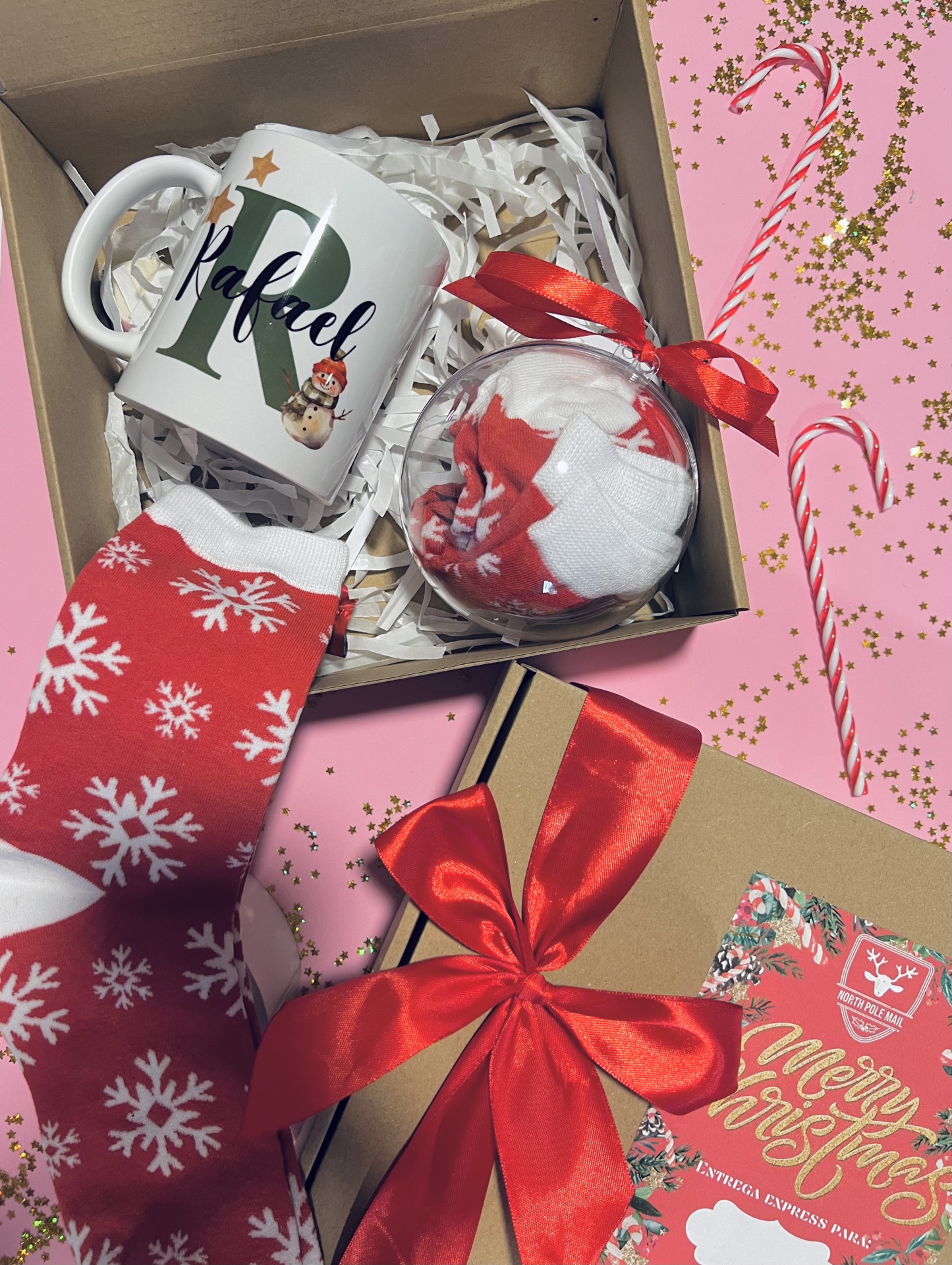 Box Caneca Inicial Natal Caixa de Natal com caneca personalizada, meias vermelhas com flocos brancos e bola transparente com mais meias, caixa de presente com laço vermelho, fundo rosa com bengalas de doces e confetes.