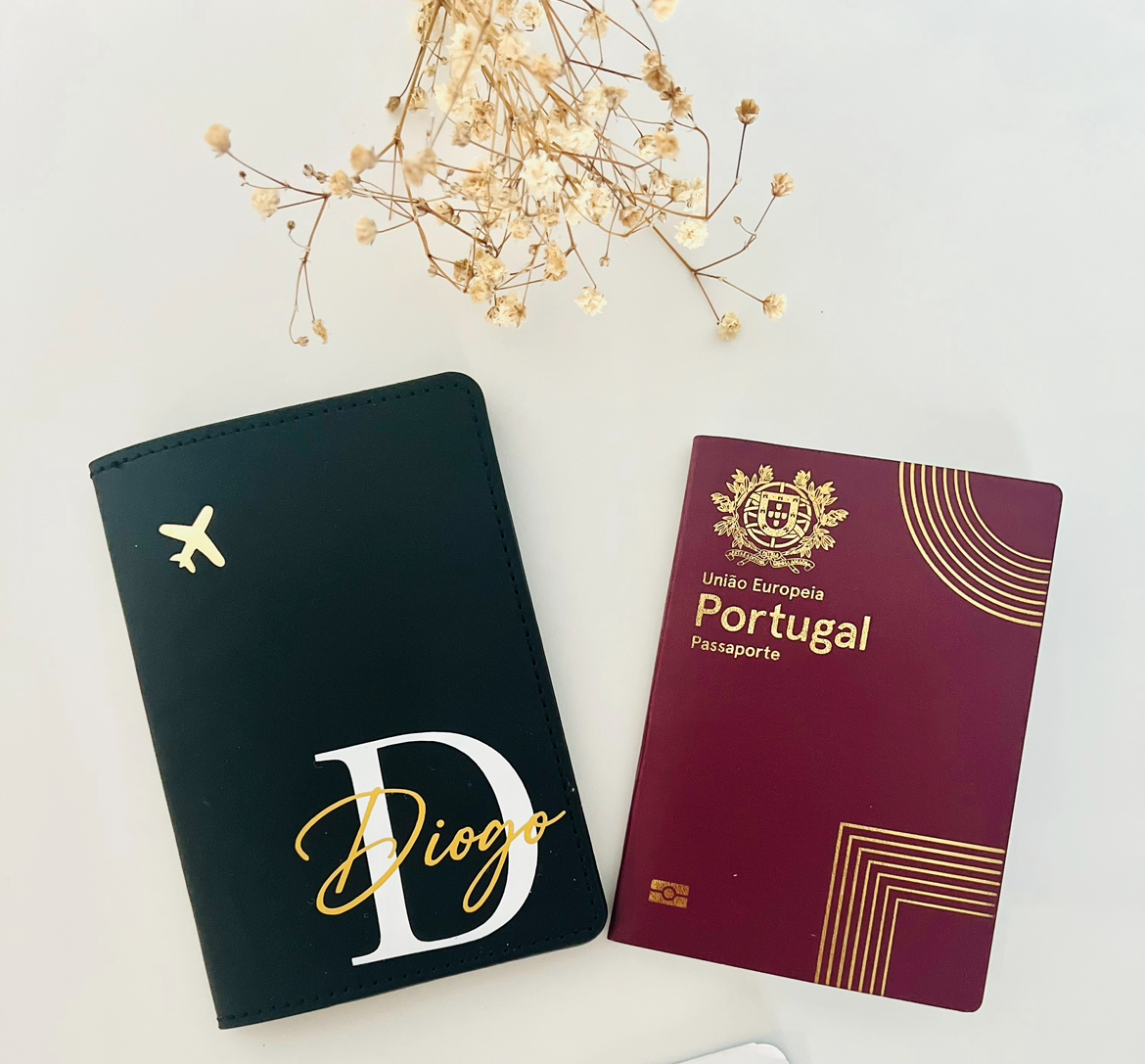 Capa de Passaporte Passaporte Portugal vinho ao lado de capa preta personalizada com avião dourado e letras, sobre fundo branco com flores secas