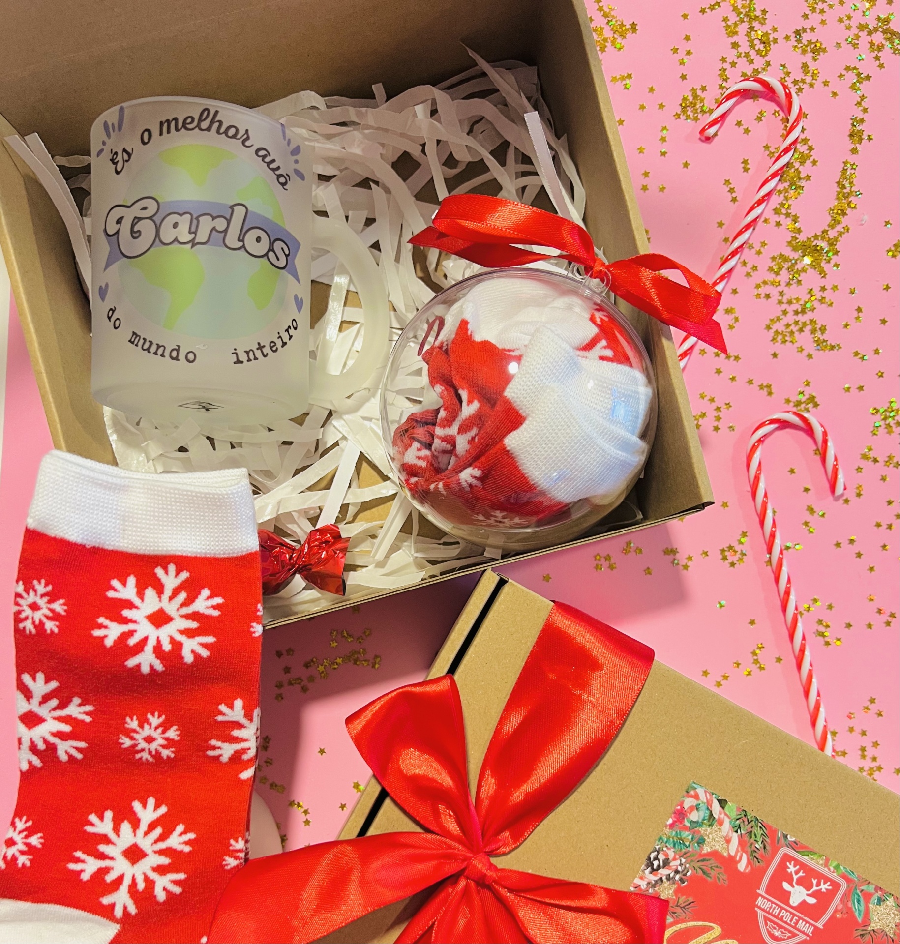 Box Caneca Mundo Caixa de presente com caneca personalizada, meias vermelhas com flocos de neve e bola de Natal, tudo decorado, sobre fundo rosa com confetes e bastões de doces.