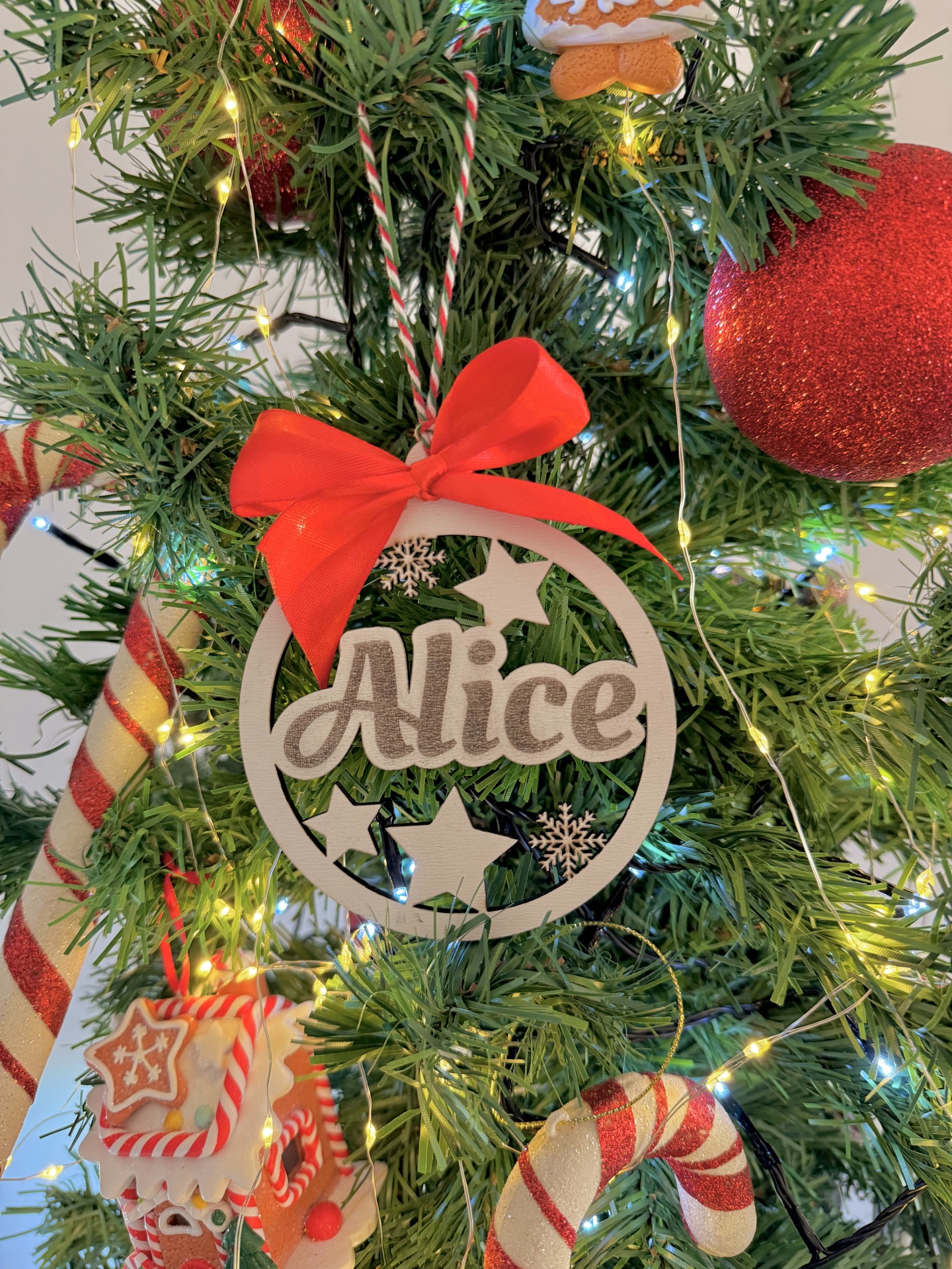 Bola de Natal com nome Decoração de Natal branca com nome Alice e laço vermelho numa árvore de Natal verde com luzes e ornamentos