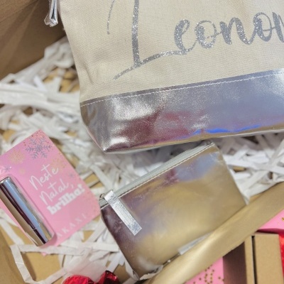 Duas bolsas com fecho, uma bege e prateada com texto 'Leonor' e outra toda prateada, com embalagem rosa e bombons numa caixa decorativa