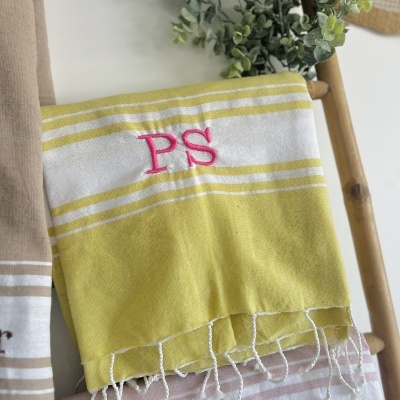 Toalhas turcas com riscas amarelas, bege e branco, bordadas com letras 'PS' e 'ML', penduradas em suporte de madeira com planta ao fundo