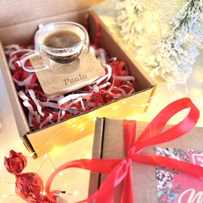 Caixa de oferta com chávena de vidro e suporte de madeira com o nome Paula, doce envolto em papel vermelho, árvores de Natal naturais com neve e luzes