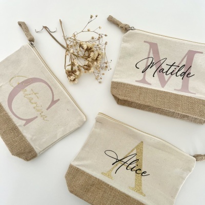 Três necessaires em tecido natural com letras e nomes personalizados e flores secas sobre fundo branco.
