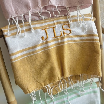 Três toalhas penduradas num cabide, rosa claro, amarelo mostarda com JLS bordado e verde menta com CL bordado.