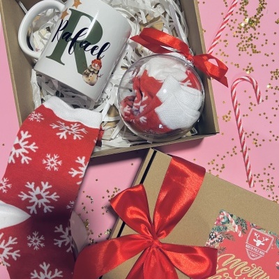 Caixa de Natal com caneca personalizada, meias vermelhas com flocos brancos e bola transparente com mais meias, caixa de presente com laço vermelho, fundo rosa com bengalas de doces e confetes.