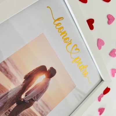 Foto em moldura branca com casal na praia ao pôr do sol e texto dourado, decoração com corações de feltro vermelhos e rosa.