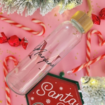 Garrafa de vidro transparente com tampa dourada e nome 'Mariana' colocada entre decoração natalícia em fundo rosa