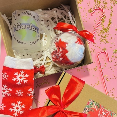 Caixa de presente com caneca personalizada, meias vermelhas com flocos de neve e bola de Natal, tudo decorado, sobre fundo rosa com confetes e bastões de doces.