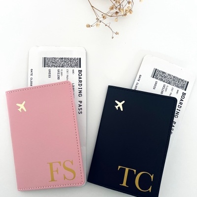 Dois porta-cartões de embarque em couro sintético rosa e azul escuro com letras douradas FS e TC e ícone de avião, com cartões de embarque, sobre fundo branco e ramo seco.