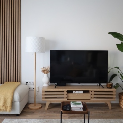 Sala de estar com móvel TV madeira clara, TV preta, candeeiro pé branco, puff cinza com manta amarelo, planta em vaso trançado