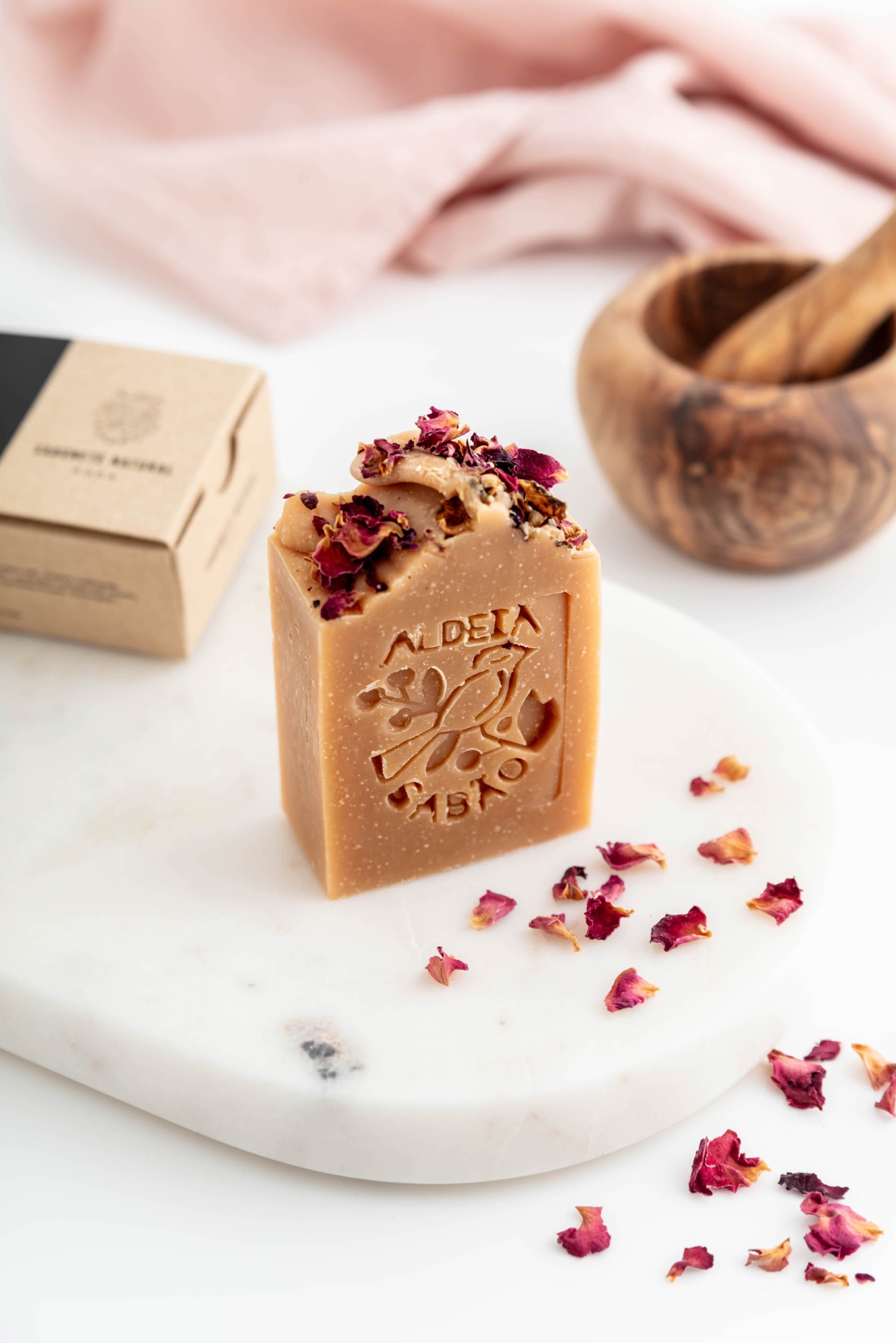 Sabonete bege com pétalas de rosa secas sobre base de mármore branco.