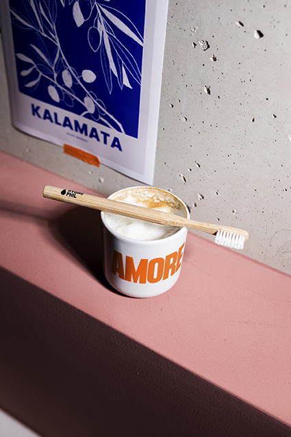 Escova de Dentes de Bambu Hydrophil - Média Escova de dentes de madeira sobre chávena com bebida e arte azul na parede.