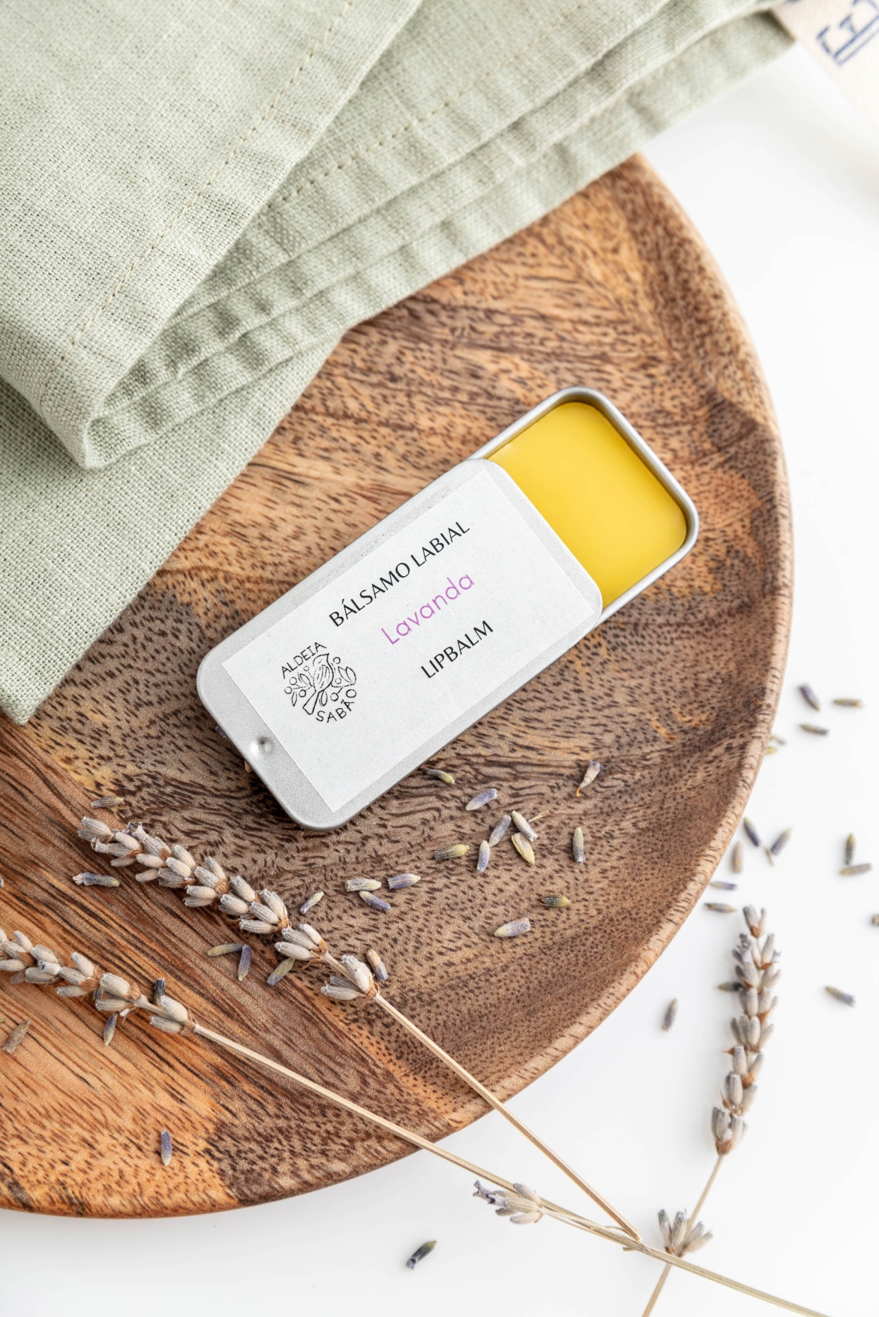 Bálsamo Labial Bálsamo labial em lata branca com texto, sobre base de madeira com lavanda seca