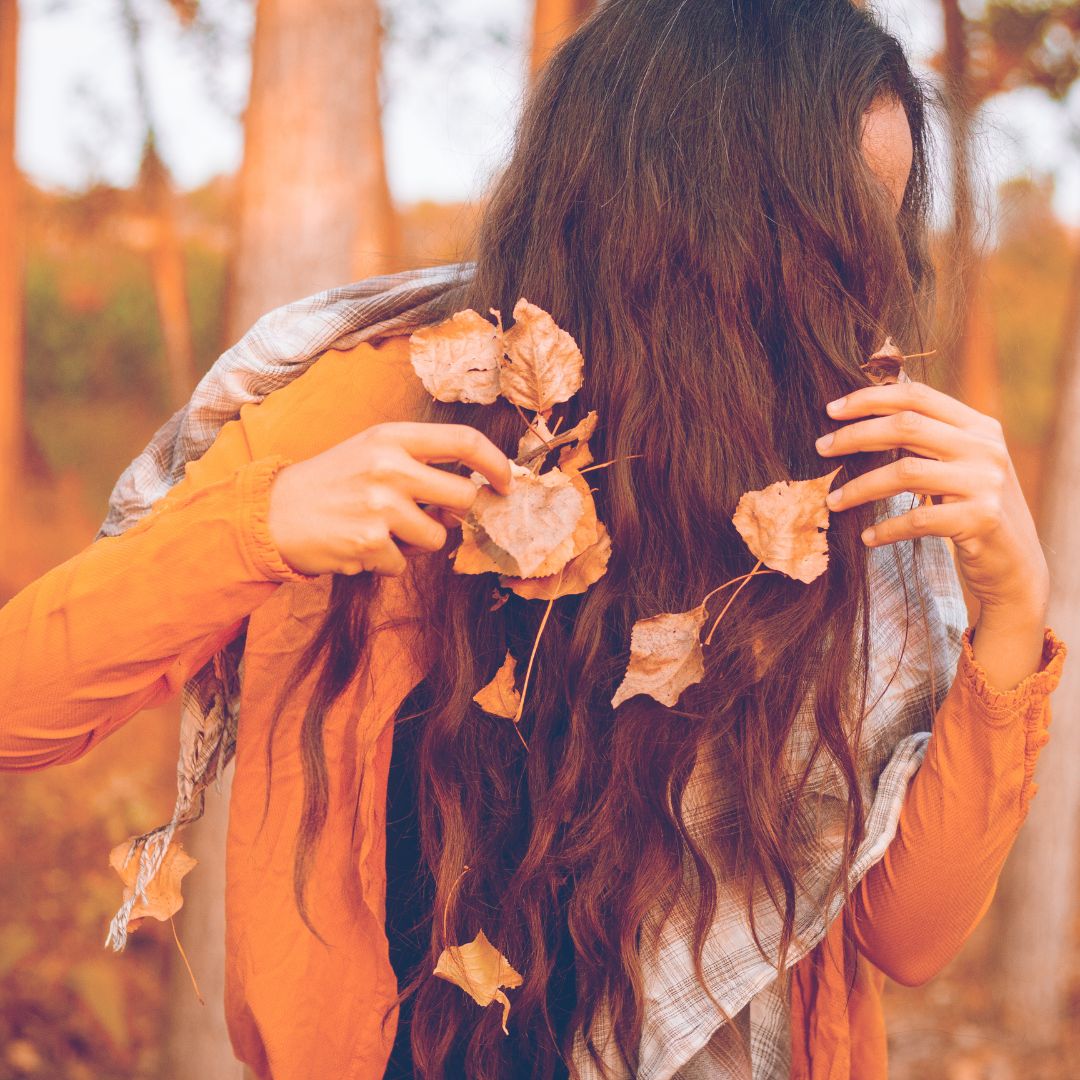 Pessoa com cabelo comprido a segurar folhas secas e vestindo camisa laranja e lenço cinzento num ambiente exterior