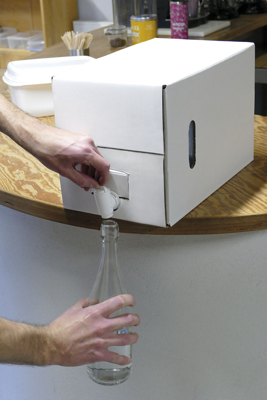 Vinagre de Limpeza (granel) Caixa dispensadora branca com torneira e garrafa de vidro numa bancada de madeira