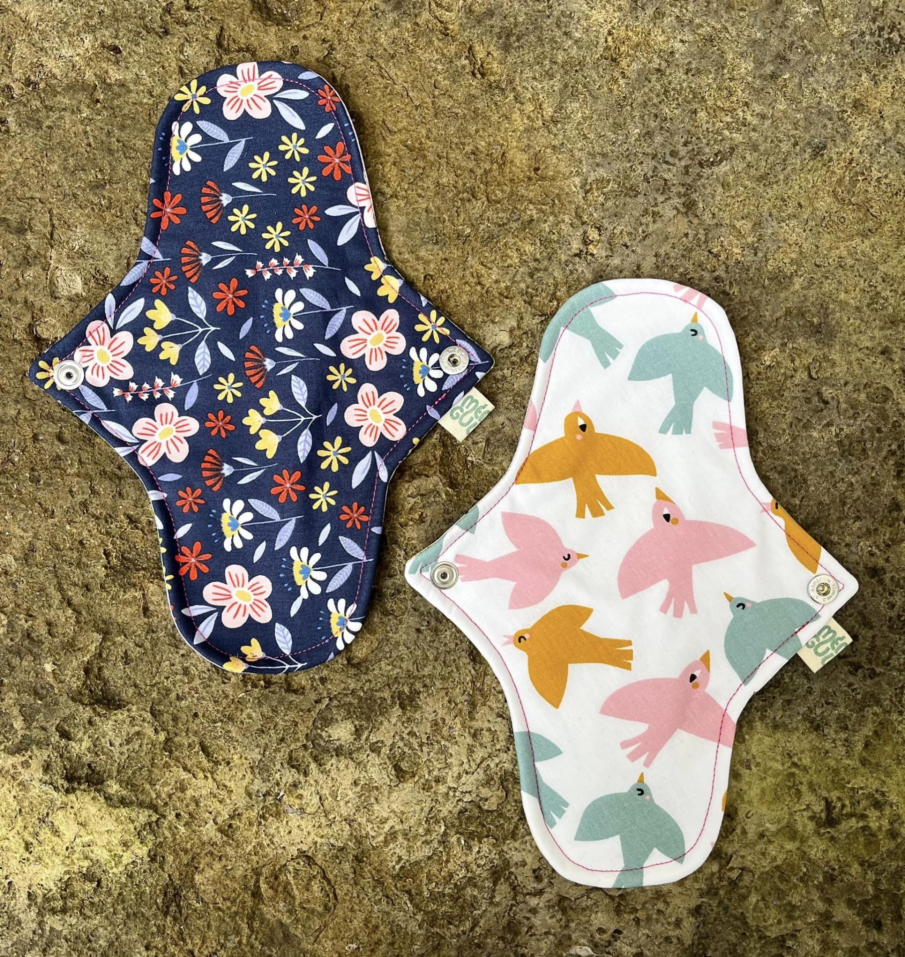 Dois pensos higiénicos reutilizáveis com padrões coloridos sobre superfície rochosa