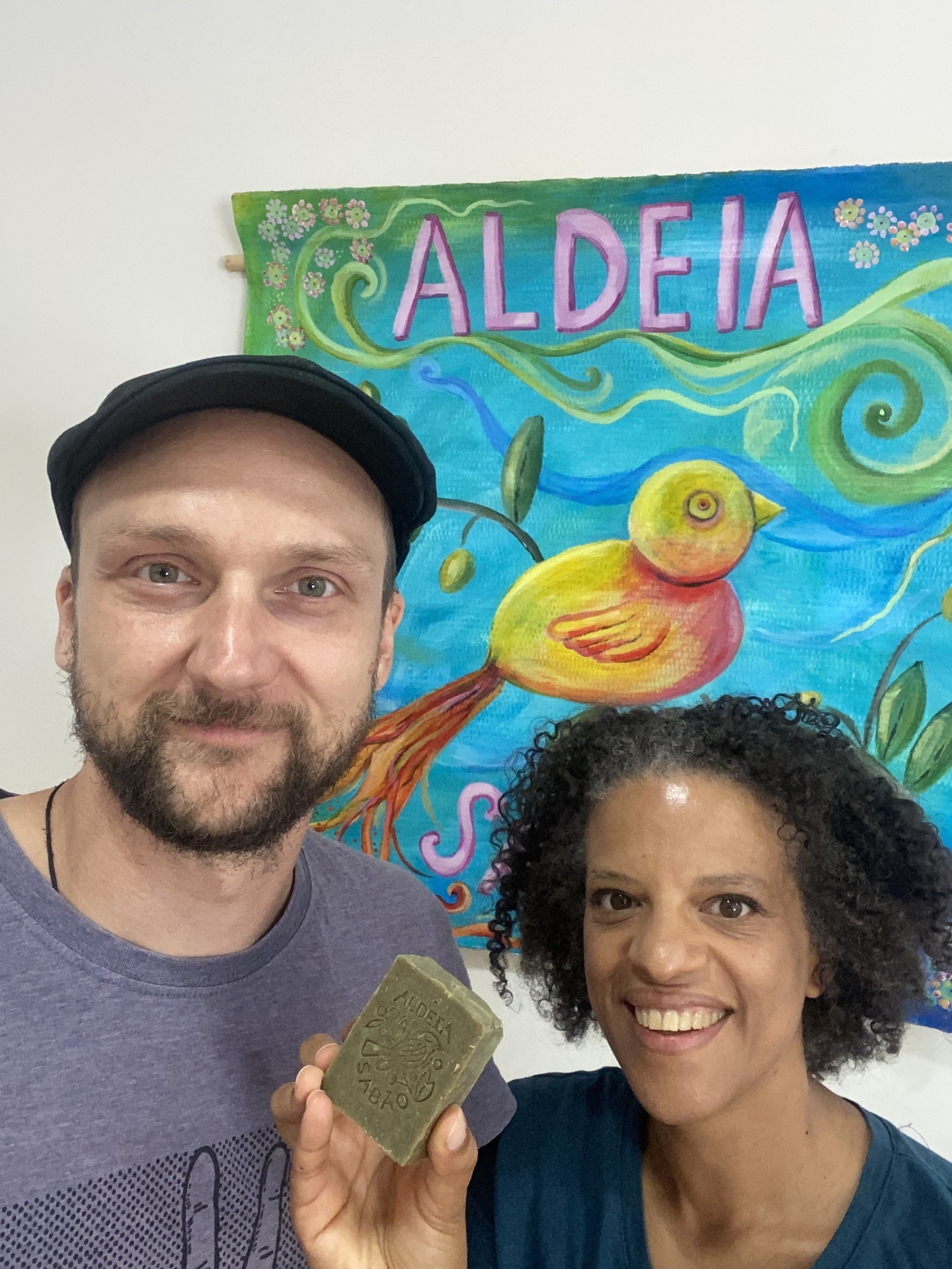 Duas pessoas seguram um sabonete artesanal verde diante de uma pintura colorida com pássaro e palavra ALDEIA no fundo.