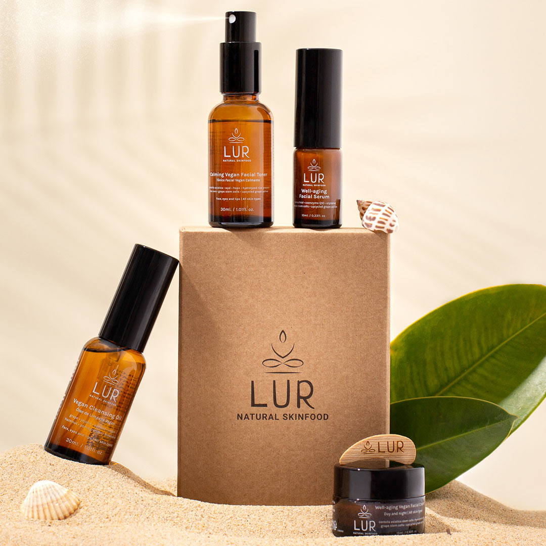 Pack 4 produtos Well-Aging (tamanho viagem) Produtos LUR Natural Skinfood em frascos âmbar e embalagem de cartão marrom sobre areia com folha verde e conchas