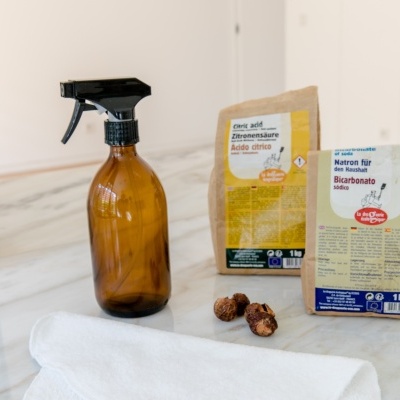 Frasco spray âmbar com saco de ácido cítrico, saco de bicarbonato, bolinhas e pano sobre superfície branca