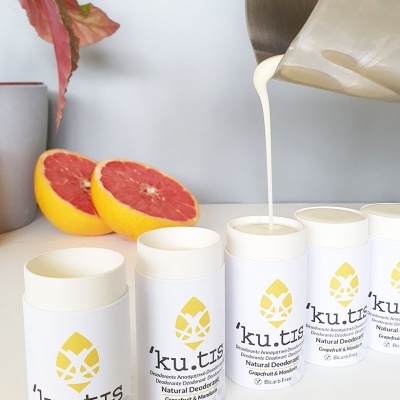 Desodorizantes naturais 'ku.tis' em embalagens brancas com detalhes amarelos, meio toranja e jarro a verter líquido cremoso