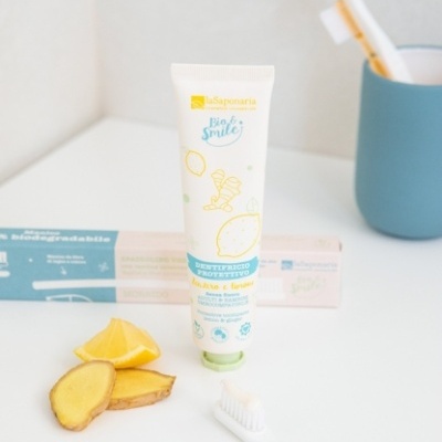 Tubo de pasta de dentes branca com tampa verde claro, escova de dentes branca, fatias de limão e gengibre sobre superfície branca