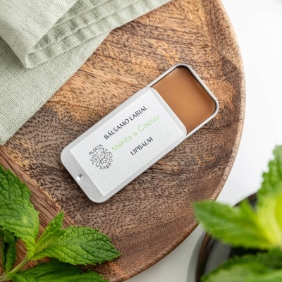 Bálsamo labial Menta & Cacau em embalagem metálica sobre tábua de madeira com folhas de hortelã