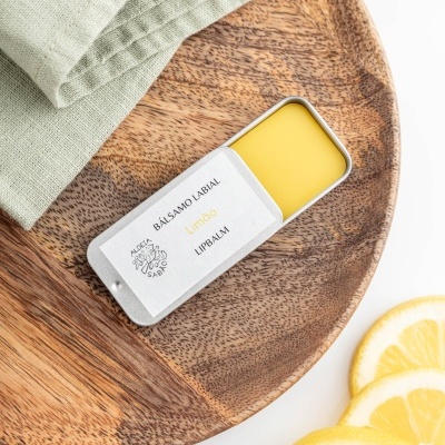 Bálsamo labial de limão em recipiente branco sobre placa de madeira com fatias de limão e tecido verde