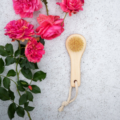 Escova facial de madeira clara com cerdas naturais ao lado de rosas cor-de-rosa com folhas verdes sobre superfície cinza