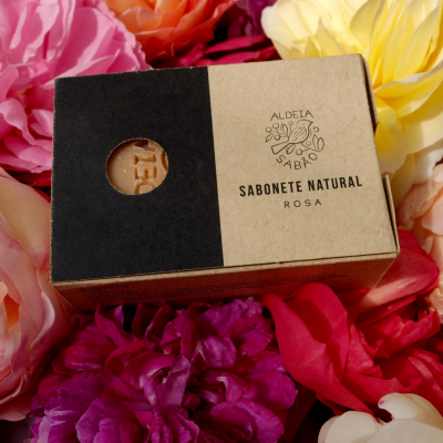Sabonete natural em embalagem de cartão com texto e abertura circular, rodeado por flores coloridas