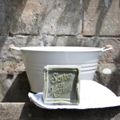 Sabonete verde quadrado 'Savon de Marseille' sobre pano branco em frente a bacia metálica branca