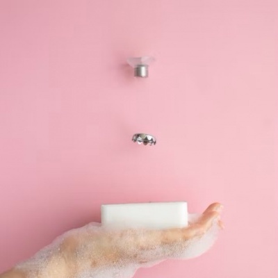 Mão com espuma segurando barra de sabão branca com torneira cromada ao fundo rosa