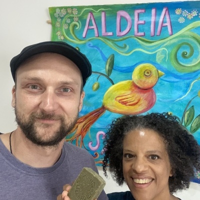 Duas pessoas seguram um sabonete artesanal verde diante de uma pintura colorida com pássaro e palavra ALDEIA no fundo.