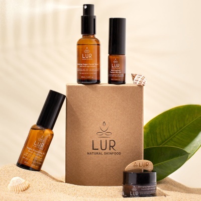 Produtos LUR Natural Skinfood em frascos âmbar e embalagem de cartão marrom sobre areia com folha verde e conchas