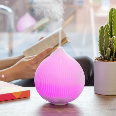 Difusor de aroma rosa iluminado em forma de gota emitindo vapor numa mesa próxima a vaso com cacto e livro aberto