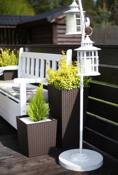 Banco de jardim branco com vasos de plantas em rattan castanho escuro e suportes de lanternas brancas num deck de madeira