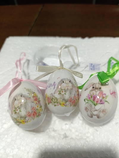 Ovos decorativos de Páscoa com coelhos e flores, com fitas rosa, creme e verde, sobre espuma branca.