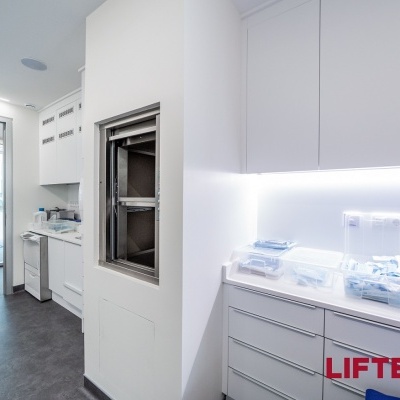 Área com armários e bancadas brancas, embalagens transparentes e porta de vidro ao fundo