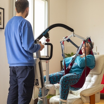 Elevador de mobilidade com assento suspenso vermelho e estrutura metálica usada por mulher assistida por homem em ambiente interior