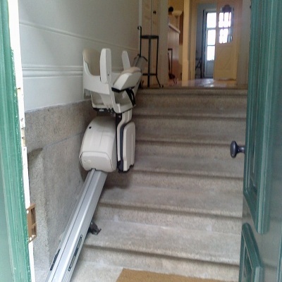 Elevador de escadas branco e preto instalado numa escada de pedra numa porta verde aberta com tapete castanho