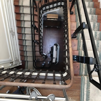 Vista superior de escada com corrimão de madeira e ferro preto, degraus com tapetes e equipamento de acessibilidade no patamar inferior.