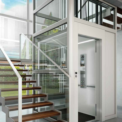 Elevador residencial moderno com estrutura metálica branca e vidros transparentes ao lado de escadas de madeira.