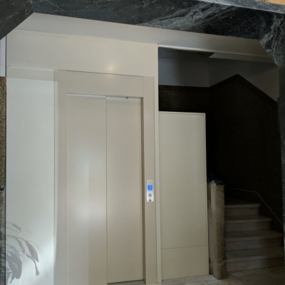 Entrada de elevador cinza claro ao lado de escada com moldura em pedra escura e texto LIFTECH Move up.