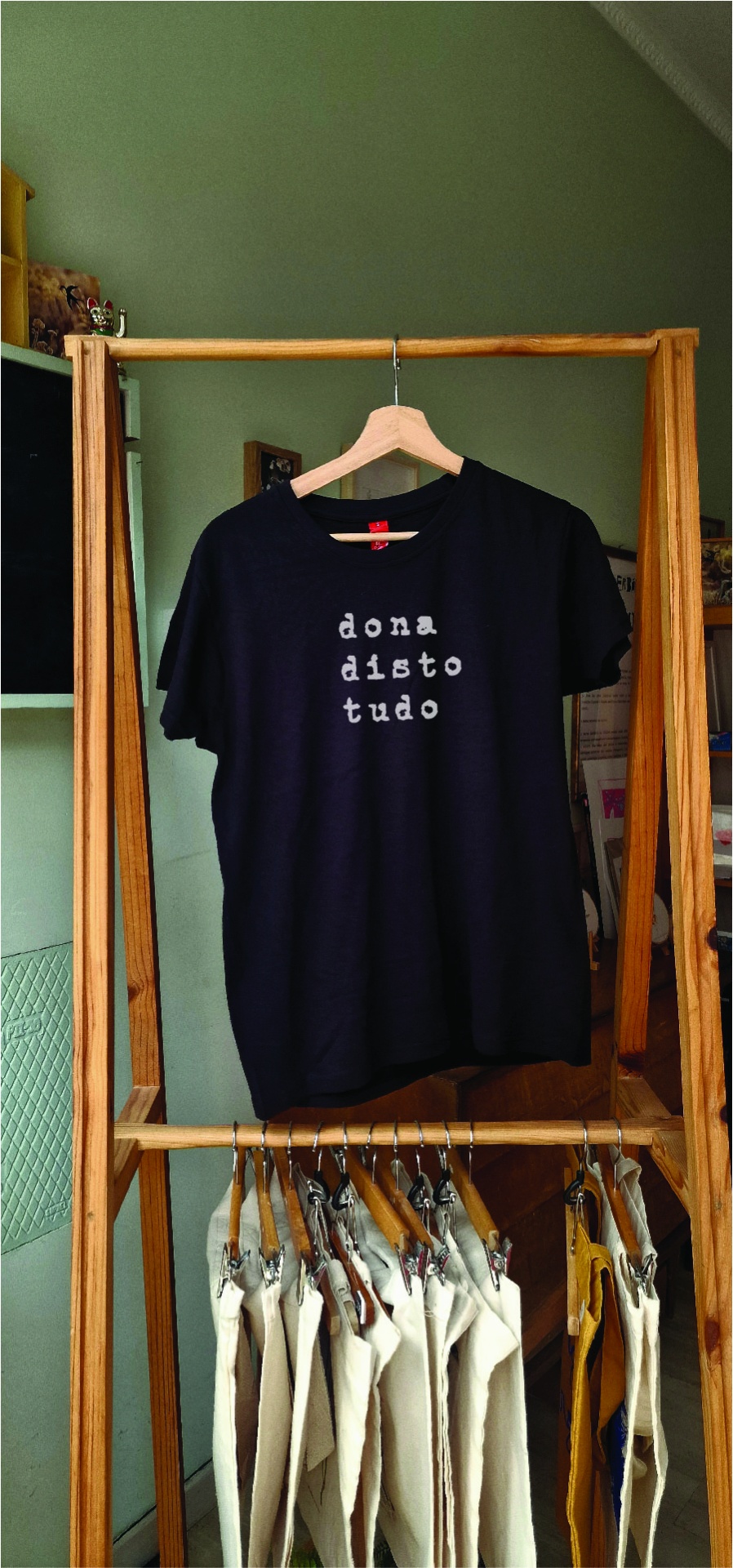 T-shirt 'dona disto tudo' em preto T-shirt preta com texto em branco pendurada numa estrutura de madeira com cabides e roupas claras penduradas