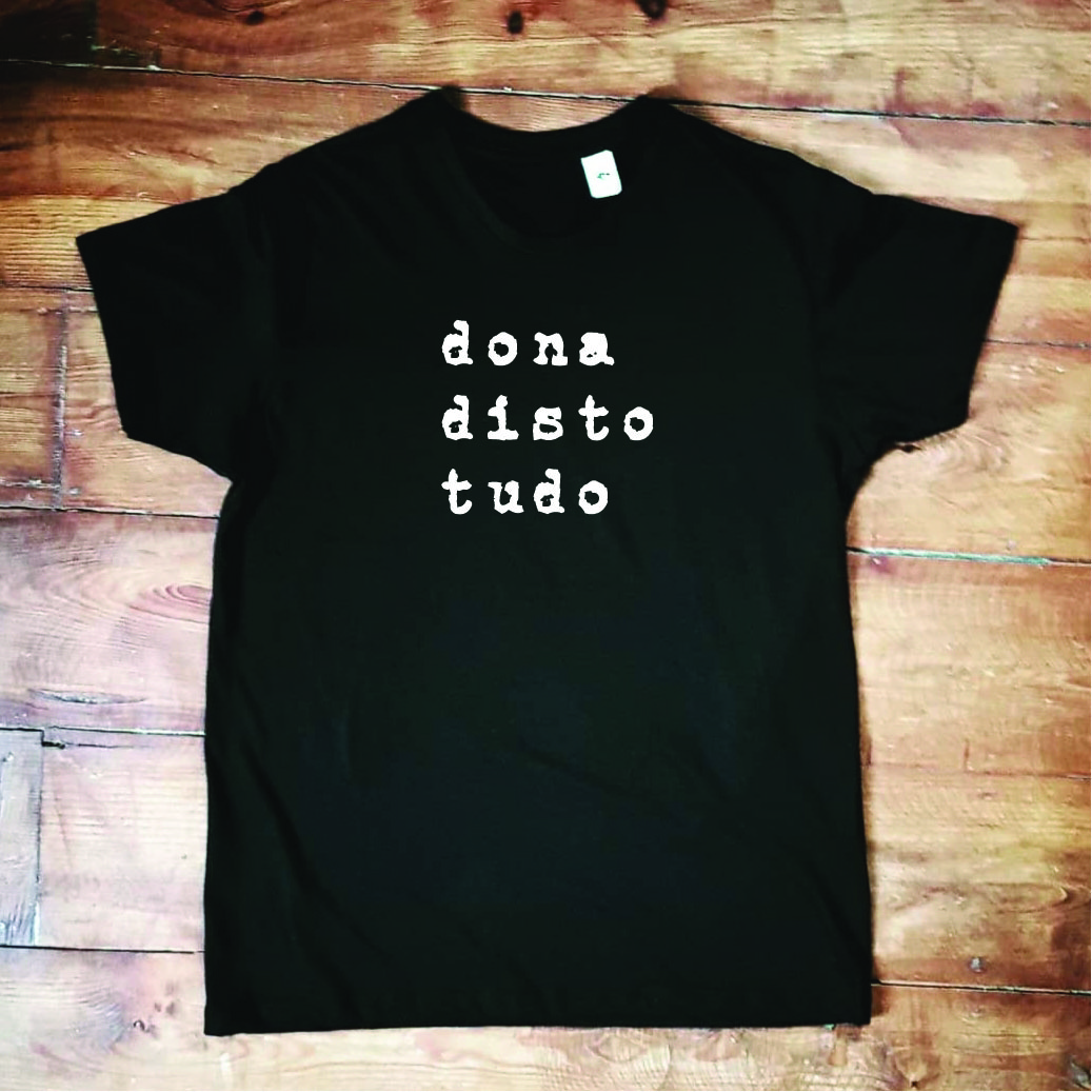 T-shirt 'dona disto tudo' em preto T-shirt preta com texto branco 'dona disto tudo' sobre chão de madeira clara