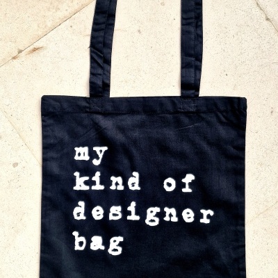 Saco tote bag preto com texto branco e alças sobre piso de pedra claro.