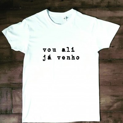 T-shirt branca com texto preto 'vou ali já venho' sobre madeira escura