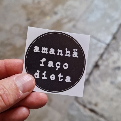 Etiqueta autocolante redonda preta com texto branco 'amanhã faço dieta' sendo segurada por uma mão