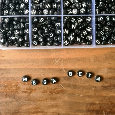 Contas pretas com letras brancas para bijuteria, organizadas em caixa de plástico e sobre mesa de madeira