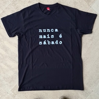 T-shirt preta com texto branco 'nunca mais é sábado' sobre chão claro com pernas e sapatos visíveis